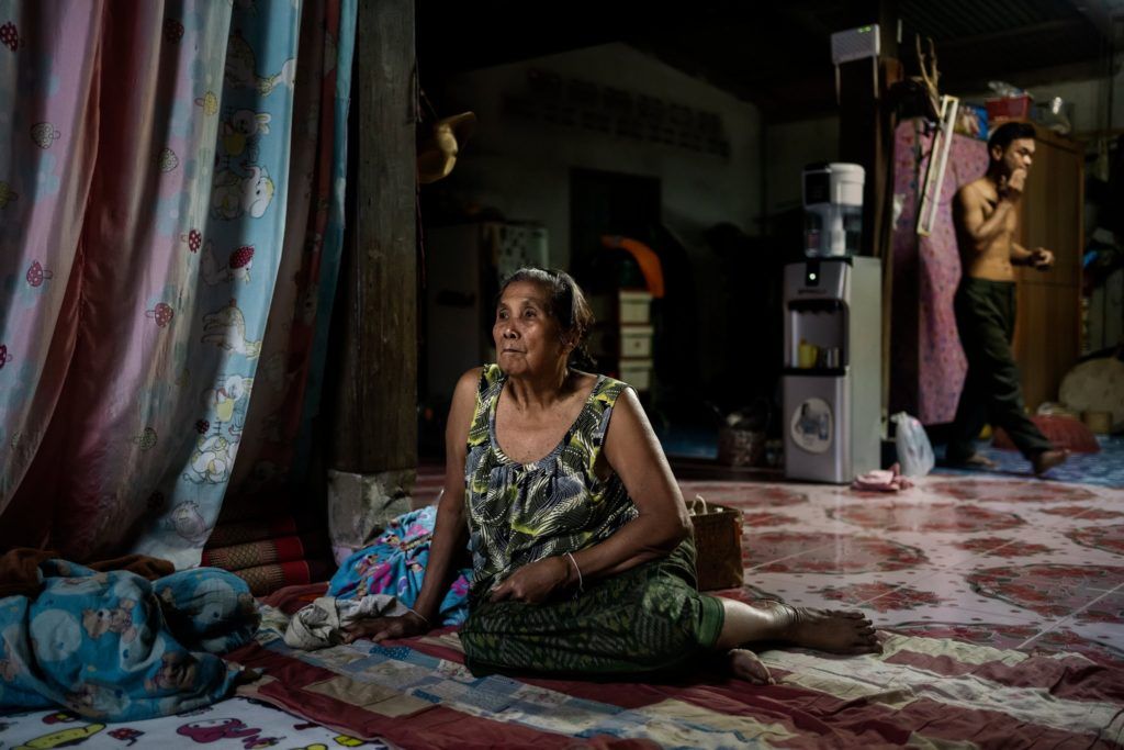 Bà Amkha Janlong xem TV trong lúc con rể, Wittaya Thongnet, đi phía sau, trong ngôi nhà họ sống bên sông Mekong. Ảnh: The New York Times.
